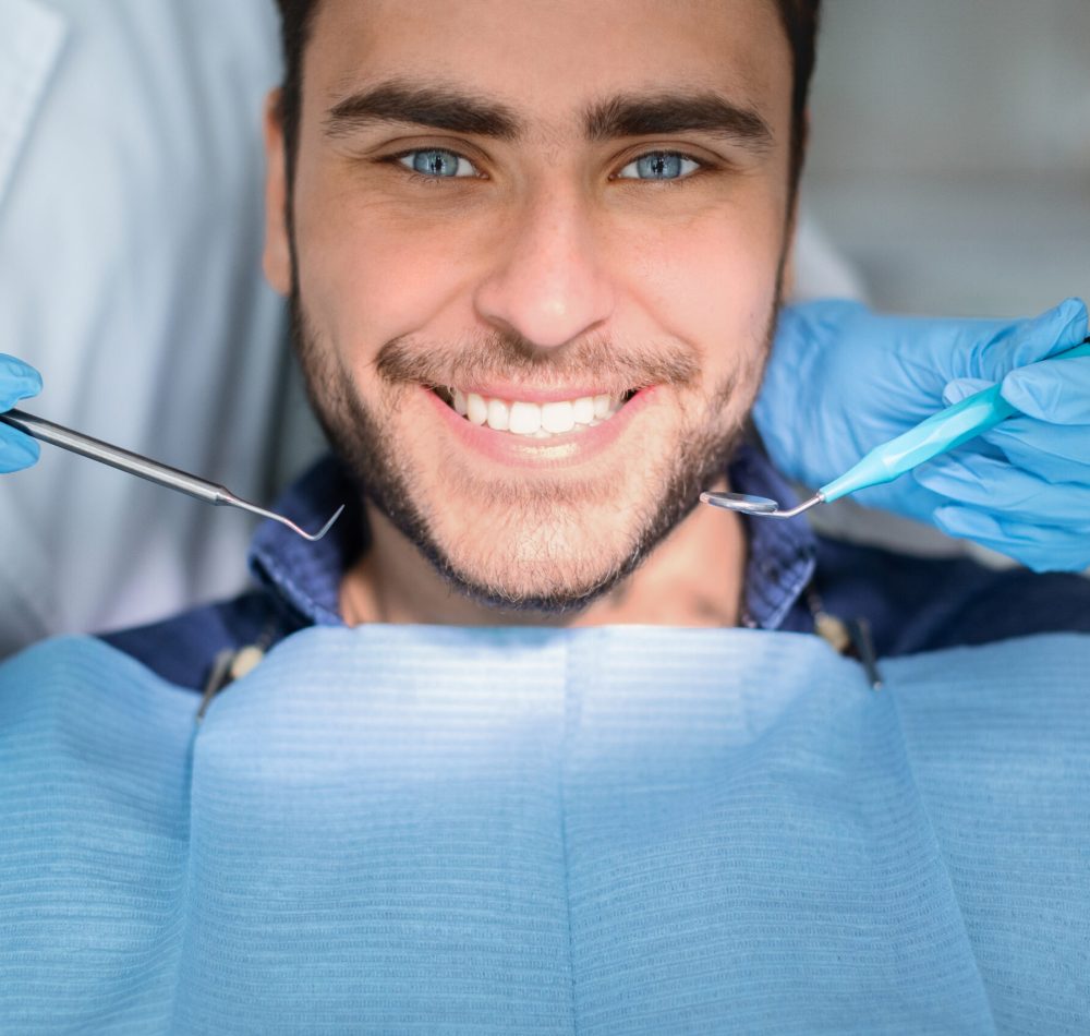 Closeup of handsome young man showing his white teeth while having checkup at dental clinic, dentist hands in rubber gloves holding dental tools. Male patient getting teeth whitening treatment