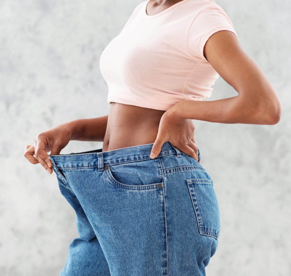 Unrecognizable black woman in oversized jeans showing results of her weight loss diet or liposuction, light background. Millennial lady achieving success in slimming, leading healthy lifestyle